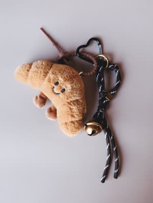 A plush croissant-shaped bag charm with an embroidered smiley face, attached to a heart-shaped carabiner clip and braided cord with a gold jingle bell — the kind of thing that makes your bag feel like it has a personality.
