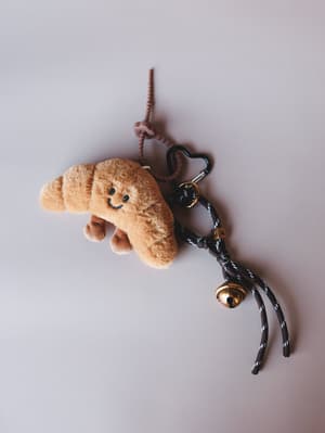 A plush croissant bag charm with an embroidered smile sits against a soft grey background, clipped to a heart-shaped carabiner with braided cord, a letter bead, and a gold jingle bell — the kind of little thing that makes your bag feel like yours.