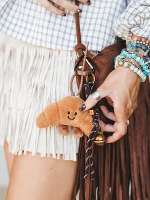 A close-up lifestyle shot of a plush croissant bag charm clipped to a brown suede fringe bag, held by a hand stacked with turquoise and stone bead bracelets — the kind of detail shot that makes you want to rearrange your whole keychain.