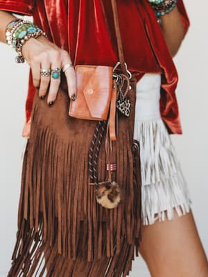A close-up detail shot of a brown suede fringe crossbody bag styled with a tan leather card holder charm, dangling charms including a leopard print heart and fur pom, against a red velvet top and white fringe skirt — the whole look dripping in layere
