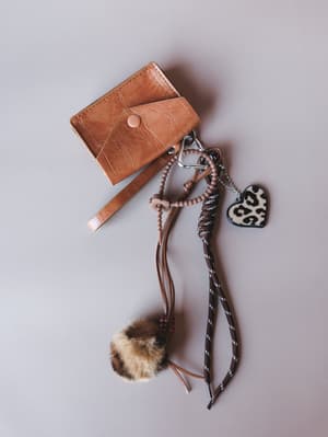 A styled flat lay of a tan croc-embossed mini card holder with a cluster of bag charms — a fluffy tortoiseshell pom, a leopard print heart pendant, and a braided cord wristlet strap — all layered together on a soft grey surface.