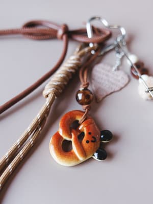 A whimsical ceramic pretzel bear bag charm rests on a soft blush surface, strung on a braided tan and brown cord with silver carabiner hardware and a blush heart charm — playful, handcrafted, and deeply emotionally attachable.