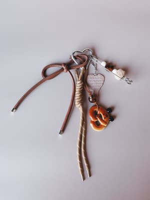 A collected little cluster of charms — heart carabiner, engraved heart pendant, pretzel charm, beaded safety pin, and knotted brown cord bow — styled flat on a soft grey surface like something you'd find in the bottom of a very cool bag.