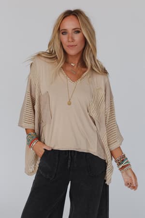 A woman with long blonde hair wears the City Drifter Oversized Contrast Top in taupe with mesh panels, paired with wide-leg pants and layered accessories for a boho chic look. She stands against a plain light background, gazing confidently at the camera.