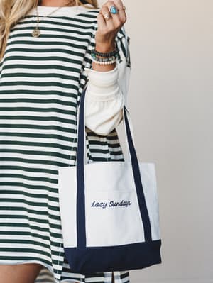A close crop of a model holding a cream canvas tote with navy trim and 'Lazy Sundays' script embroidery, styled against a striped dress and stacked boho bracelets — the kind of bag that makes a farmers market run feel intentional.