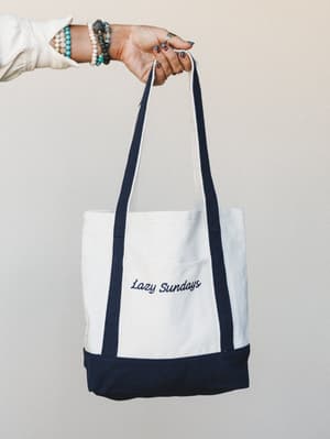 A hand holds up a structured canvas tote with navy trim and a 'Lazy Sundays' embroidered script — styled with stacked beaded bracelets and a ring, giving it that effortlessly personal, everyday-carry energy.