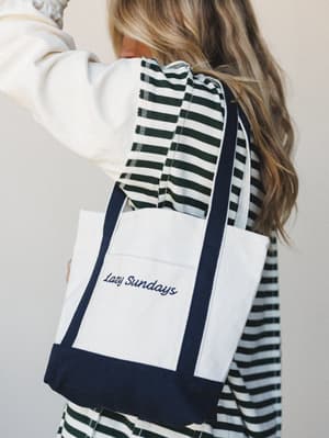 A canvas tote bag with navy trim and embroidered 'Lazy Sundays' script is slung over the shoulder of a model in a cream sweatshirt and dark green striped pants — the whole vibe is relaxed, intentional, and ready for a slow morning out.