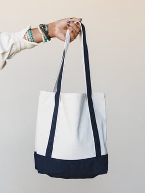A hand holding up a structured canvas tote with navy color-block trim and straps against a soft neutral wall — the stacked bracelets and blue nail polish do half the styling work.