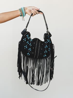 A hand holds up a black suede fringe hobo bag covered in turquoise and silver studs, styled with a stack of beaded bracelets — the kind of bag that does the talking for you.