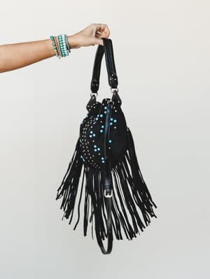 A hand holds up a black fringe hobo bag covered in silver and turquoise studs, stacked beaded bracelets on the wrist adding an effortless boho finish against a soft neutral backdrop.