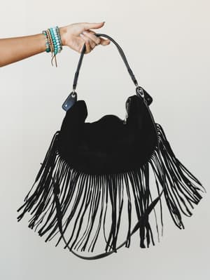 A hand holding up a black suede fringe hobo bag against a soft neutral backdrop — the long, dramatic fringe and stacked turquoise bracelets do all the talking.