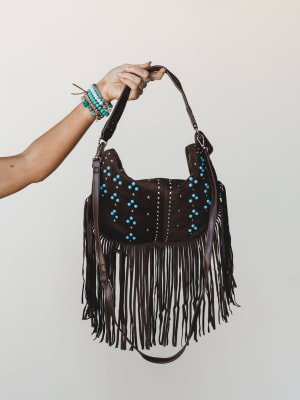 A hand holds up a rich chocolate suede hobo bag dripping with long fringe and turquoise studding — styled with a stack of beaded bracelets that feel like they belong together.