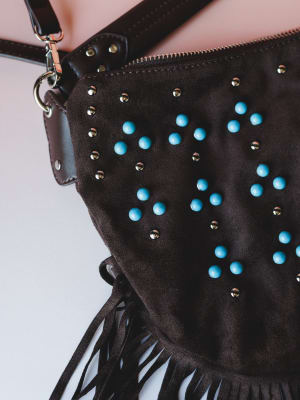 A close-up detail shot of a chocolate suede hobo bag covered in turquoise dome studs and gold mini studs, with leather fringe along the bottom edge and a crossbody strap — the kind of bag that feels like it belongs on a road trip through the Southwes