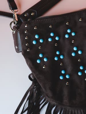 A close-up detail shot of a chocolate suede hobo bag covered in turquoise dome studs and gold mini studs, with leather fringe along the bottom edge and a crossbody strap — the kind of bag that feels like it belongs on a road trip through the Southwes