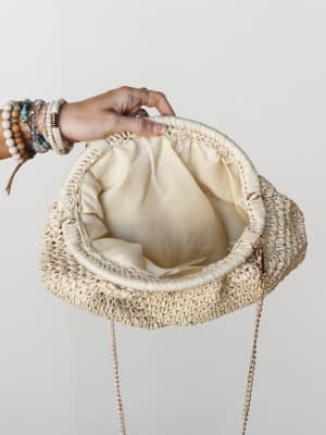 A hand adorned with stacked boho bracelets and rings holds open a woven raffia clutch, revealing its cream fabric lining and gold chain strap — the kind of bag that makes a sundress feel intentional.