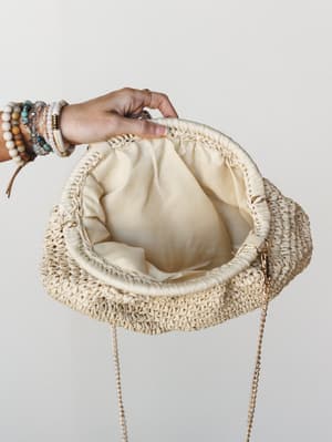 A hand adorned with stacked boho bracelets and rings holds open a woven raffia clutch, revealing its cream fabric lining and gold chain strap — the kind of bag that makes a sundress feel intentional.