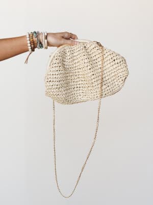 A hand holds up a chunky crochet raffia clutch in warm ivory, its gold chain strap cascading down — styled with a stack of beaded and wood bracelets that feel like a summer collected over years.