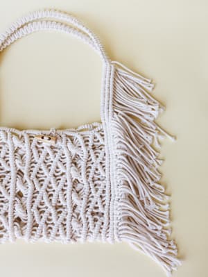 A close-up of a macrame tote bag in soft ivory, shot flat against a warm cream background — the knotted texture and fringe trim do all the talking.