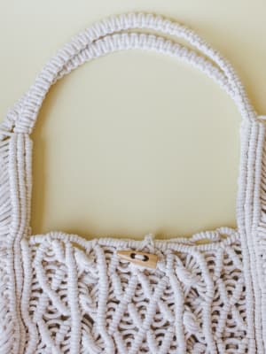 A close-up detail shot of a handcrafted macramé tote in soft ivory, with knotted rope texture, a single looped top handle, and a small wooden toggle button closure — the kind of bag that makes a farmers market run feel like a whole moment.
