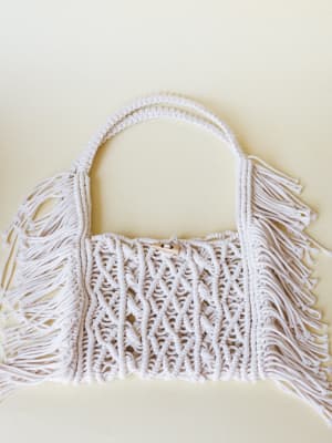 A cream macramé handbag with fringe sides and a single top handle, laid flat against a soft ivory background — the kind of bag that makes a sundress feel intentional.