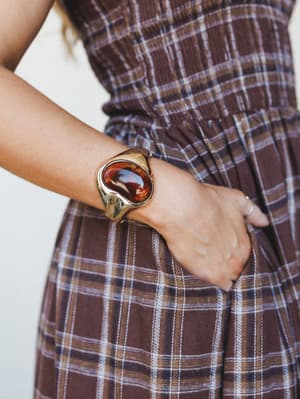 A close-up wrist shot spotlighting a sculptural gold cuff bracelet with a rich amber stone centerpiece, worn against a brown plaid dress — the kind of accessory that quietly anchors a whole outfit.