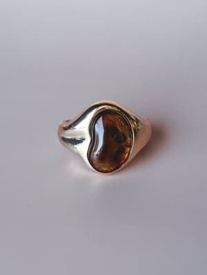 A polished gold signet-style ring with an oval amber cabochon stone, shot clean against a soft grey surface — the kind of piece that looks like it's been in someone's jewelry box for twenty years and just got rediscovered.
