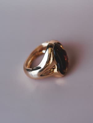 A chunky gold ring with a dark amber stone sits against a soft lavender-grey surface — the kind of piece that looks like it's been in someone's jewelry box for twenty years in the best way.
