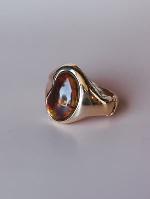 A chunky gold signet-style ring with a smooth oval amber-brown cabochon stone, shot close up against a soft lavender-grey surface — the kind of piece that looks like it's been passed down but somehow feels completely current.
