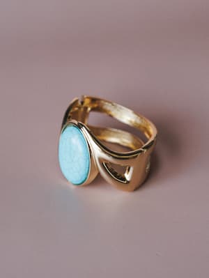 A gold-toned bangle bracelet with a large oval turquoise stone sits on a soft mauve surface, giving off that collected, well-traveled energy — the kind of piece that feels like it came home from somewhere interesting.