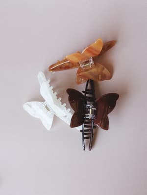 Three butterfly-shaped claw clips arranged in a casual overlap on a soft blush background — one amber tortoise, one pearlescent white, one deep brown — the kind of hair accessories that make a ponytail feel intentional.
