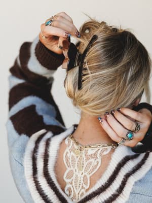 Shot from behind, this close-up captures the layered details that make an outfit feel intentional — a striped fuzzy sweater, lace bralette peeking through the back, gold layered necklaces, turquoise and silver rings stacked on both hands, leopard nai