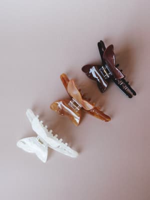 Three butterfly claw clips arranged diagonally on a soft blush background — one pearlescent white, one warm tortoiseshell amber, one deep burgundy — the kind of everyday accessories that quietly pull a whole look together.