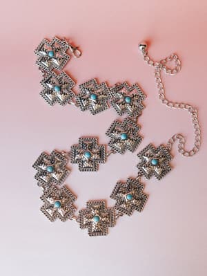 A silver concho chain belt laid flat on a blush pink surface — each square medallion is etched with a cross motif and centered with a turquoise stone, connected by rope-twist chain links with an adjustable extender.