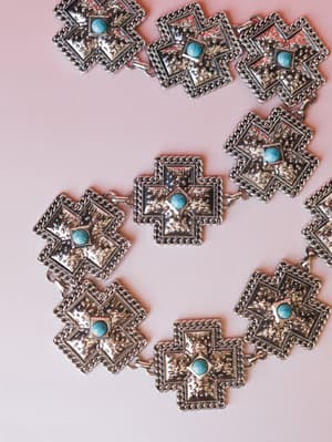 A silver concho chain belt laid flat on a blush pink surface — each hammered square link centered with a turquoise stone and edged in rope-style chain detail, giving it that southwestern-meets-boho energy.