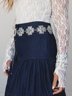 A mid-shot detail of a white lace long-sleeve top tucked into a dark indigo denim tiered skirt, anchored by a statement silver chain belt with cross-shaped concho links and turquoise stone accents — the kind of outfit that does a lot of talking on it