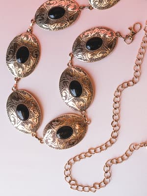 A gold concho chain belt with oval engraved medallions and black stone centers, styled flat on a soft blush background — the kind of accessory that makes a simple dress feel like a whole personality.