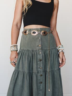 A mid-shot of a tiered denim midi skirt in washed olive paired with a black crop top, styled with a gold concho chain belt featuring black stone medallions — the whole look layered with stacked beaded bracelets and rings for that effortless boho-west