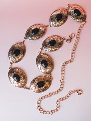 A gold concho chain belt laid flat on a soft pink surface — oval engraved medallions with black stone centers linked by a chunky gold chain, equal parts western and boho.