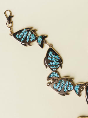 A silver-toned link bracelet or belt made of connected fish-shaped charms, each inlaid with crushed turquoise stone chips, laid out against a soft cream background — the kind of piece that looks like it was found at a coastal market and worn every da