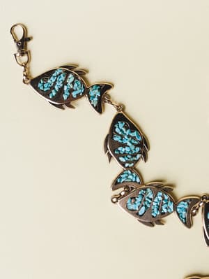 A silver-toned link bracelet or belt made of connected fish-shaped charms, each inlaid with crushed turquoise stone chips, laid out against a soft cream background — the kind of piece that looks like it was found at a coastal market and worn every da