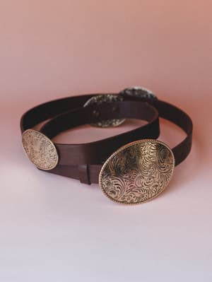 A dark brown leather belt coiled against a dusty rose backdrop, its multiple oversized oval gold conchos etched with intricate scrollwork — the kind of accessory that makes a simple outfit feel intentional.