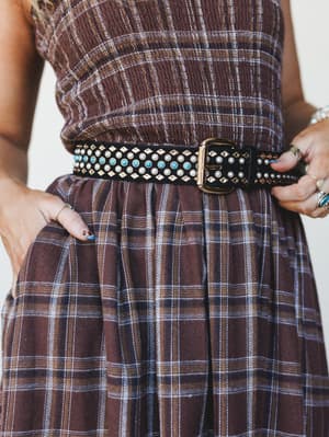 A close-up of a black studded belt cinched over a brown plaid dress — the belt does all the talking with pearl studs, turquoise stones, gold diamond hardware, and a chunky gold buckle layered over relaxed, gathered fabric.