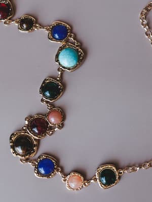 A gold chain belt laid flat, strung with round and oval cabochon stones in a rich mix of jewel tones — cobalt blue, turquoise, deep green, burgundy, blush pink, and amber — each set in an ornate gold bezel frame.