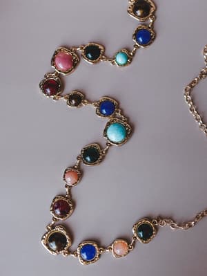 A gold chain belt laid flat, strung with round cabochon stones in a jewel-toned mix of turquoise, cobalt blue, deep red, forest green, blush pink, and smoky brown — the kind of piece that looks like it has a whole story behind it.