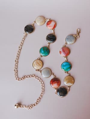 A gold chain belt with linked multicolor marbled disc medallions — each one a little different, like tiny planets — laid flat on a soft blush surface with an adjustable lobster clasp closure.