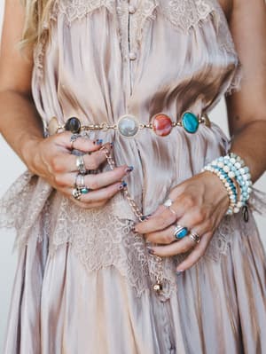 A close-up torso shot showing a satin-look mauve dress with lace trim, styled with a gold chain belt featuring colorful round stone cabochons — hands stacked with silver rings and a beaded bracelet bring the whole boho-jewelry moment together.