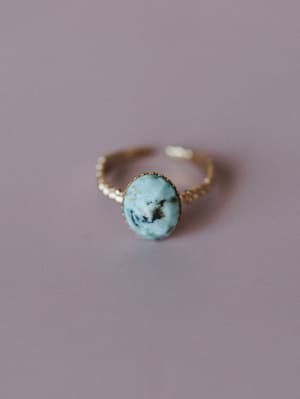 A close-up of a delicate gold open-band ring with an oval turquoise stone set in a scalloped bezel, shot on a soft lavender surface — the kind of ring you reach for every single day without thinking.