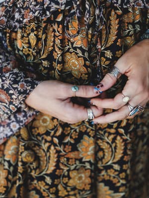 A close-up of stacked hands resting against a rich black and marigold floral print dress, layered with an assortment of silver rings and a turquoise stone ring — the kind of detail shot that makes you want to reorganize your jewelry box.