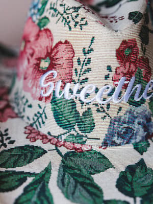 A close-up of a tapestry-style floral cowgirl hat with 'Sweethe—' embroidered in white script across the front panel — the kind of hat that has a whole personality before you even put it on.