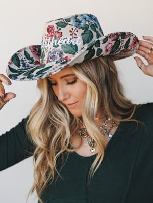 A blonde model tilts a wide-brim floral tapestry cowgirl hat downward, eyes cast low, layered gold charm necklaces catching the light against a dark forest green top — the whole thing feels like a country concert you actually want to go to.